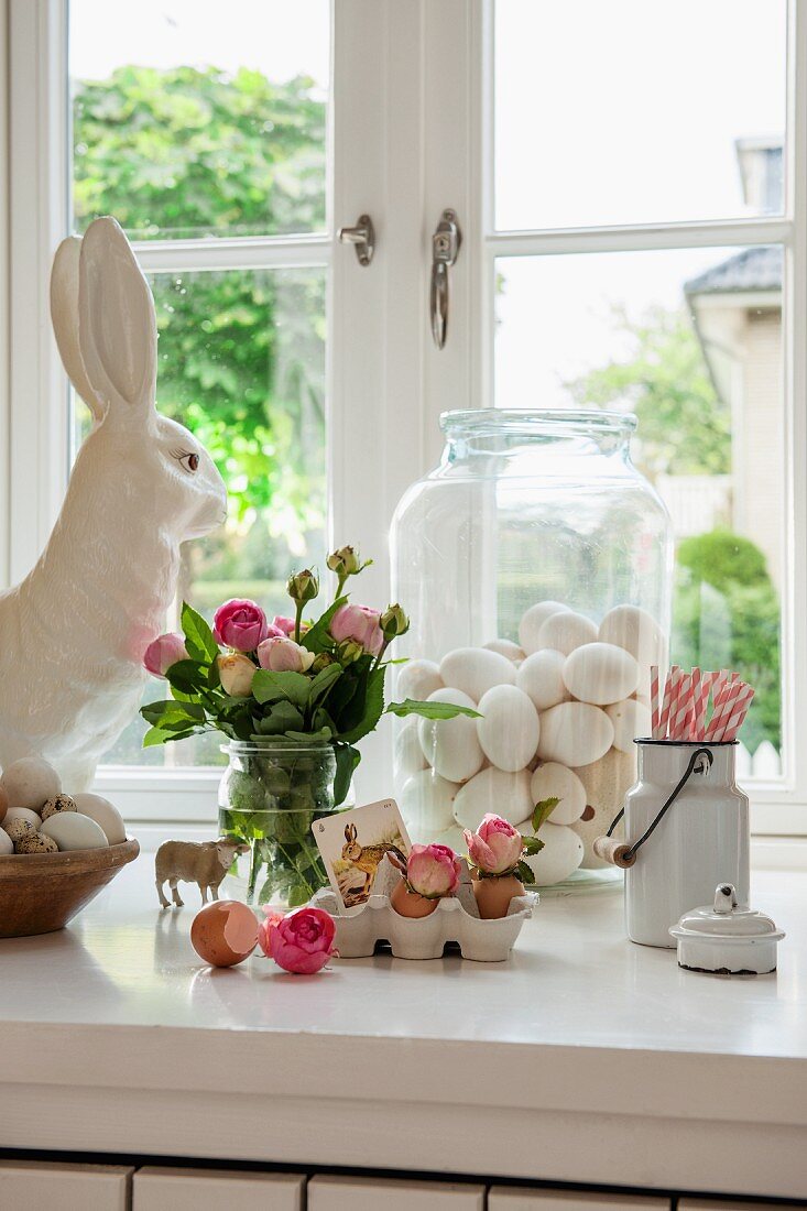 Romantische Osterdekoration auf weißer Fensterbank; ausgeblasene Eier in Glasbehälter und rosafarbener Rosenstrauss neben Porzellanhasen