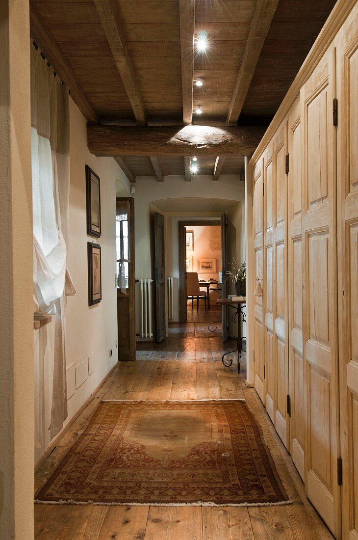 Pale, wooden custom cupboards and modern spotlights on wood-beamed ceiling in hallway