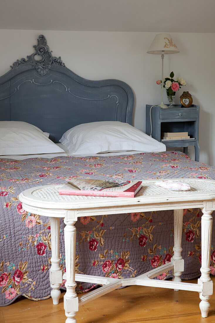 Antique side table with cane top painted white at foot of double bed with curved headboard and floral quilt