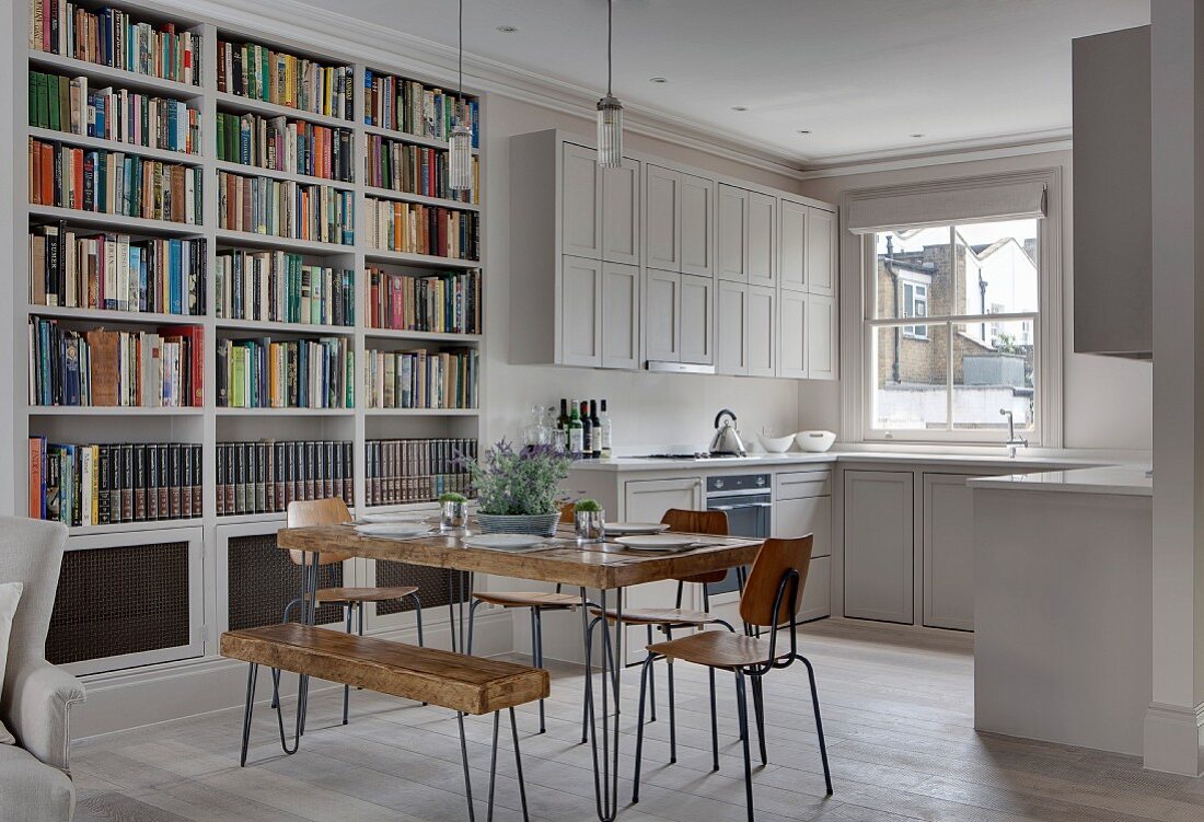 Square rustic wooden table, vintage chairs and bookcase in front of white fitted kitchen with panelled doors