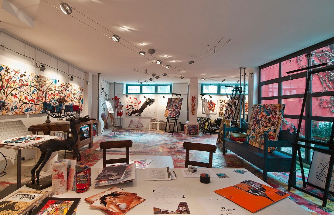 Maleratelier in Loft mit bunter moderner Malerei und Bildern an den Wänden, vorne ein großer Arbeitstisch