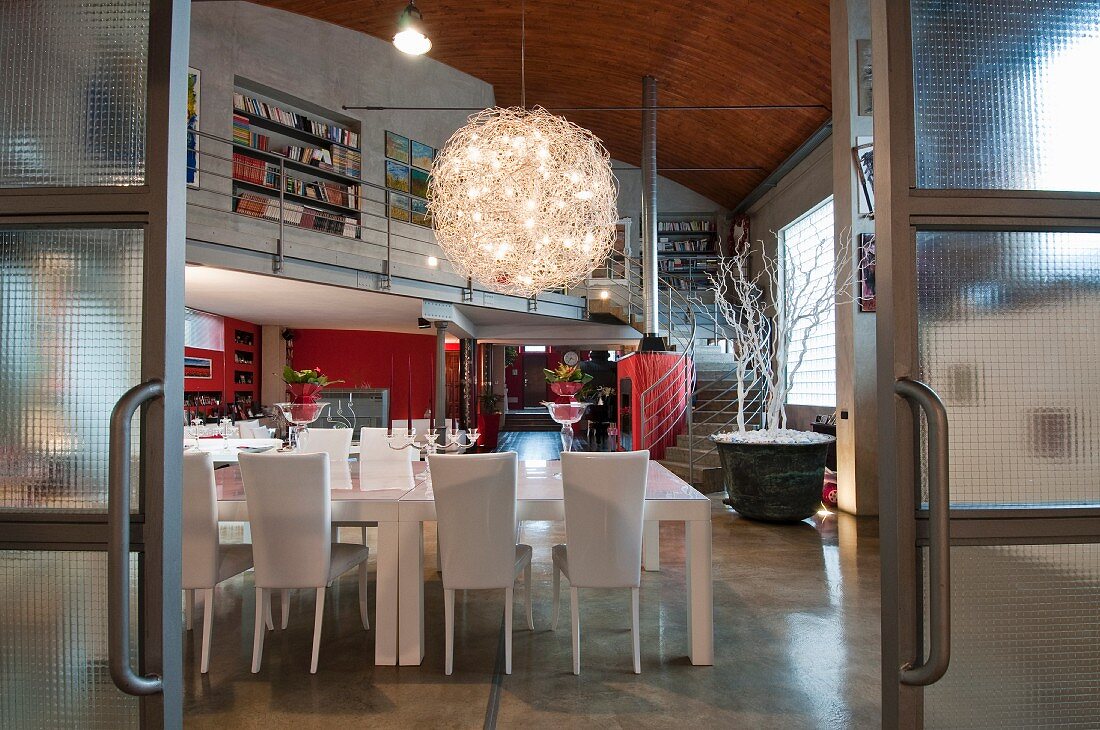 View through industrial-style glass sliding doors of white dining set below spherical chandelier in loft apartment