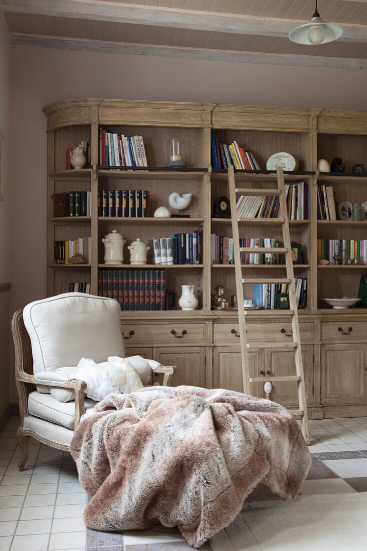 Gemütlicher Sessel mit Decke vor Bücherschrank mit Bibliotheksleiter in Landhausambiente