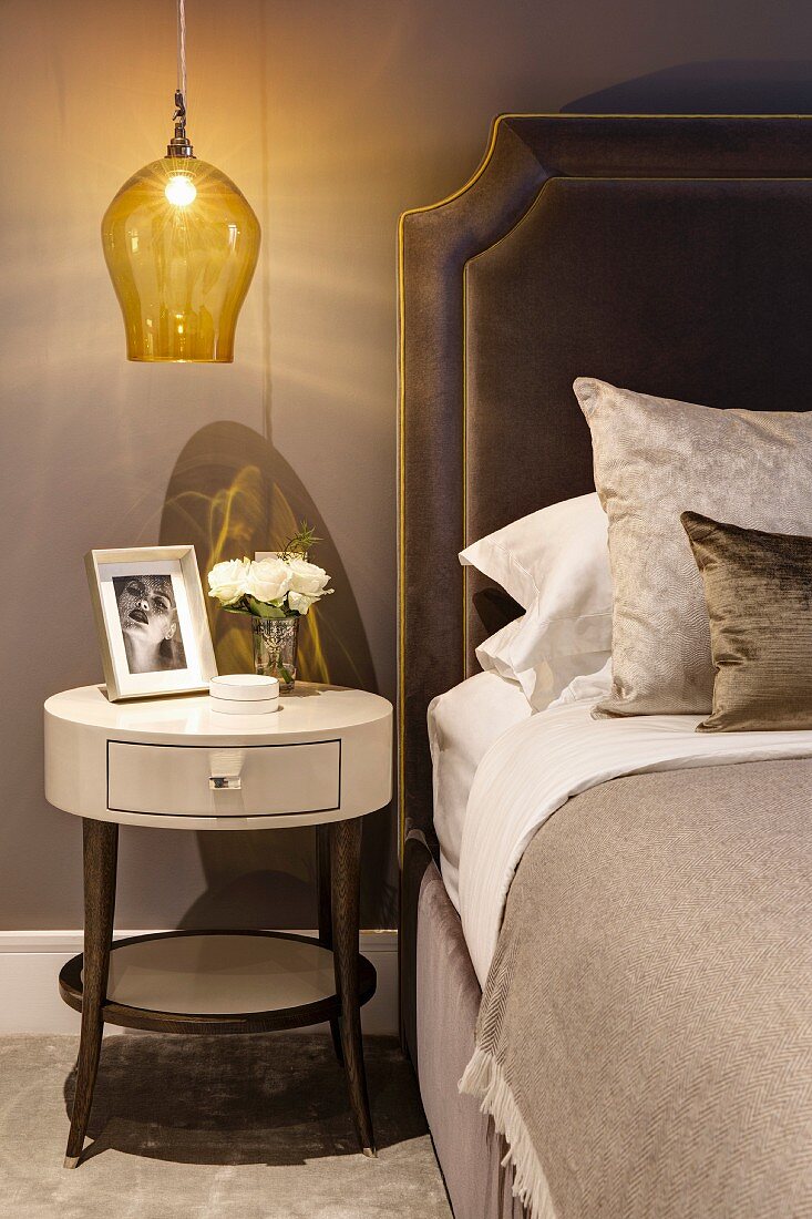 Round bedside table below pendant lamp with yellow glass lampshade next to bed with upholstered headboard