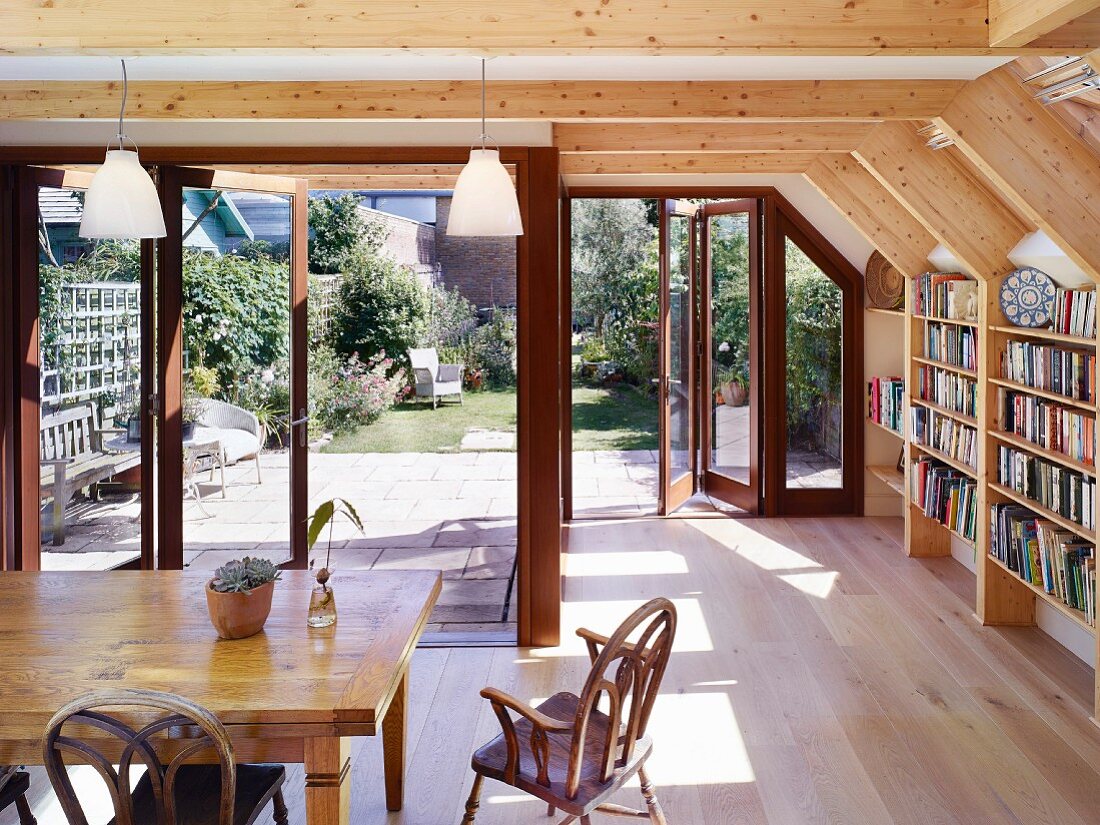 Dining area in front of open terrace doors with garden view; bookcase built into exposed wooden structure to one side