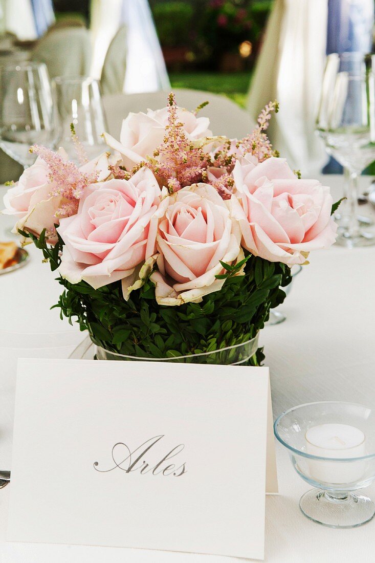 Namensschild vor Blumengesteck aus rosa Rosen, auf festlich dekoriertem Tisch