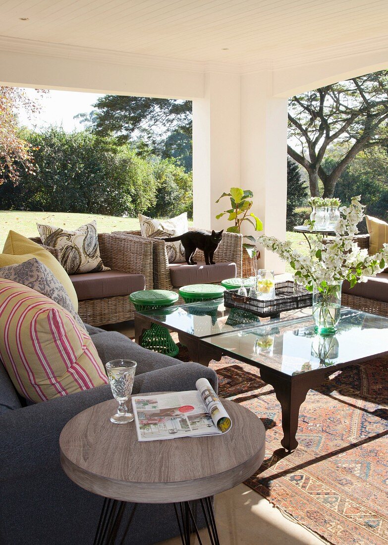 Sitzbereich auf sonniger, überdachter Terrasse mit Sofa, Rattansesseln und Coffeetable auf Orientteppich, vorne ein Beistellhocker