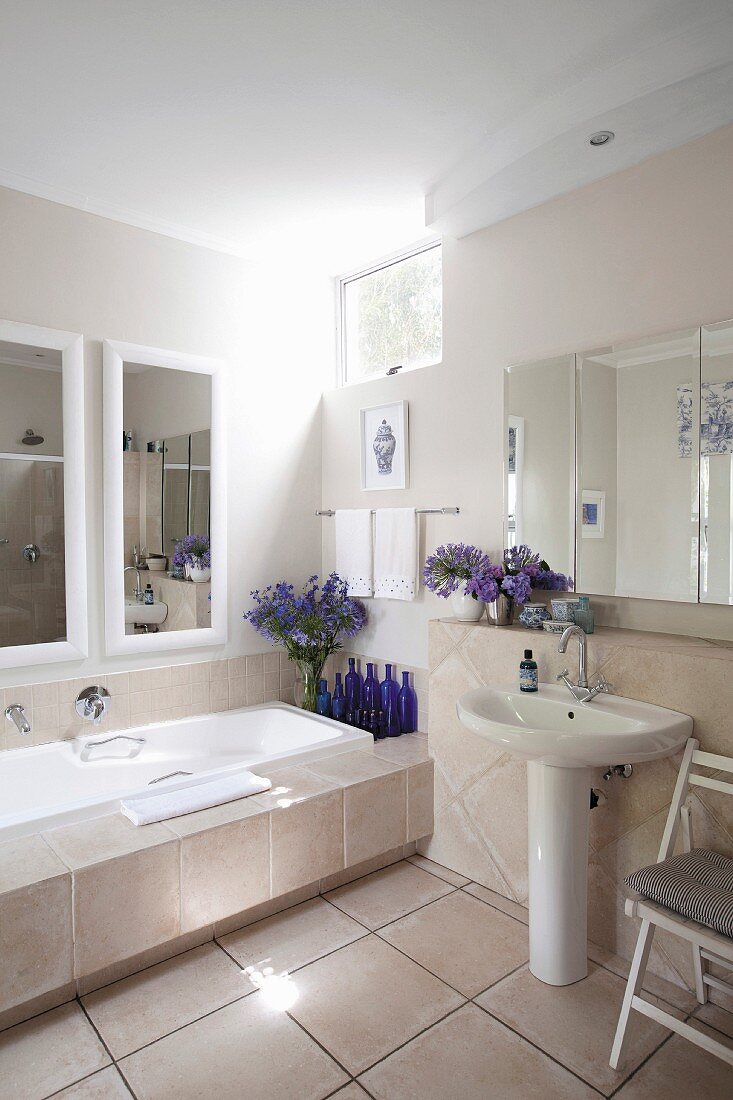 Badewanne mit breiter, gefliesten Ablage, Wandspiegel, blaue Flaschen und Blumensträusse