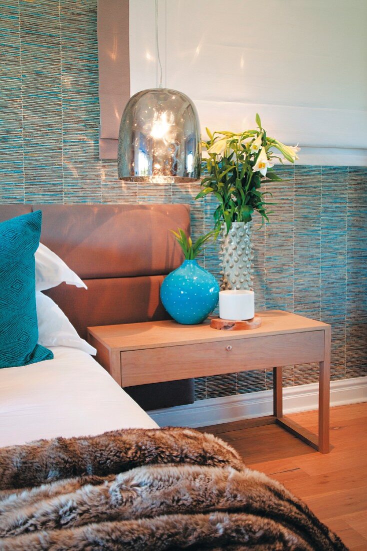 Glänzender Lampenschirm über Nachttisch und Bett mit hellbraunem Lederkopfteil vor tapezierter Wand