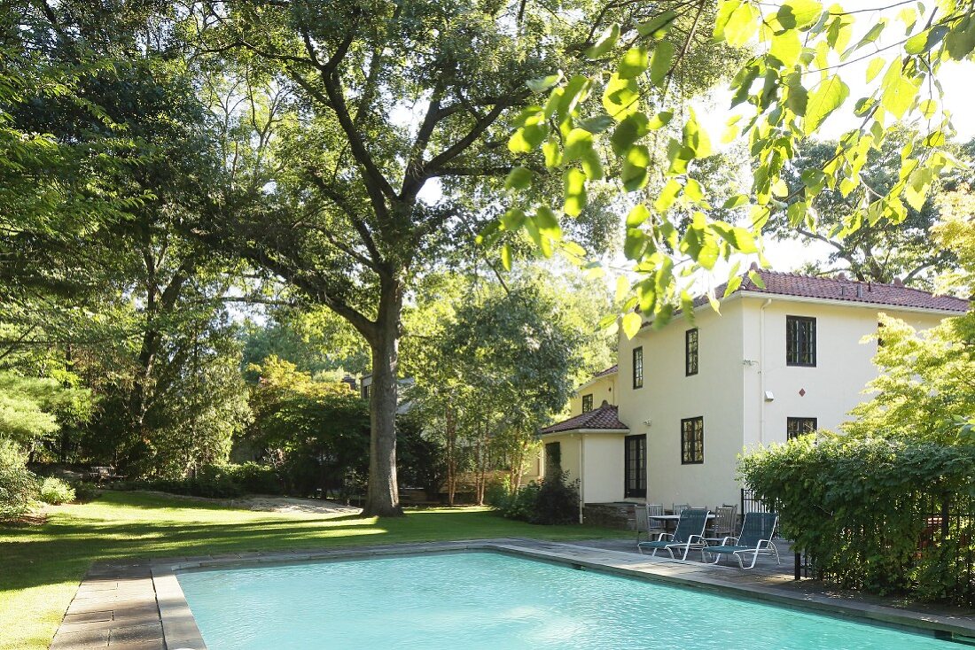 Türkisfarbener Pool in sommerlichem Garten, im Hintergrund mehrstöckige Villa