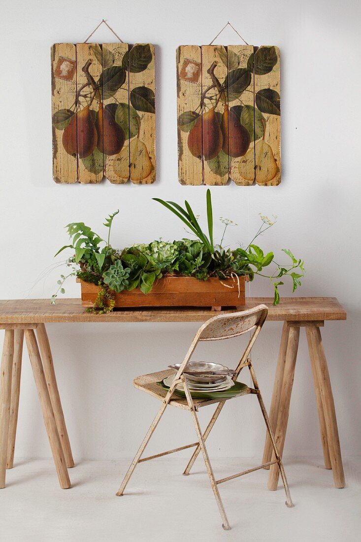 Verschiedene Grünpflanzen in Holz- Blumenkasten auf rustikalen Tisch vor aufgehängten Holztafeln mit Pflanzenmotiven