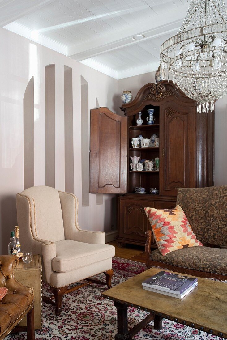 Traditionelle Wohnzimmerecke mit hellem Ohrensessel, antikem Schrank mit offener Tür und Blick auf Geschirr