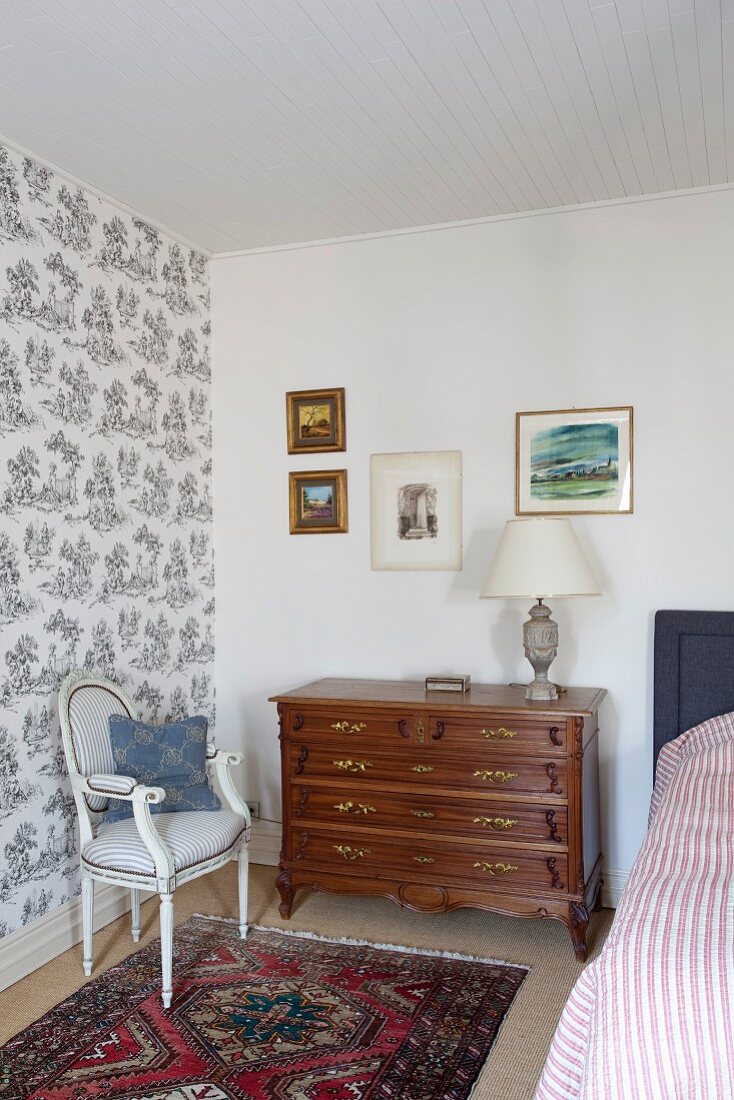 Antique chest of drawers and Rococo-style, elegant upholstered chair against wall with patterned wallpaper in corner of traditional bedroom