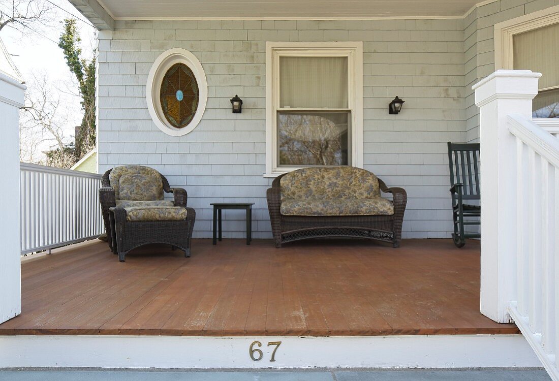 Sessel und Sofa aus dunklem Rattan mit Sitzpolstern auf Veranda vor heller Holzfassade