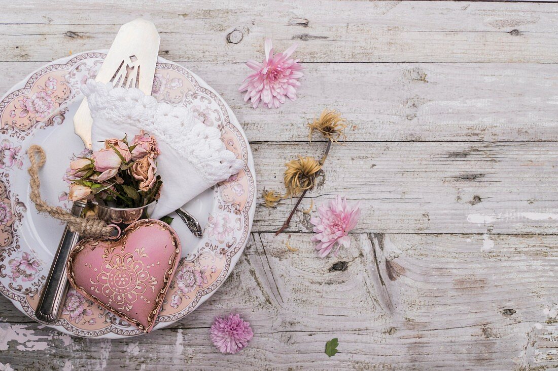 Rosa Dekoherz und getrocknetes Rosensträuschen auf nostalgischem Teller mit verstreuten Blumenblüten auf rustikaler Holzunterlage