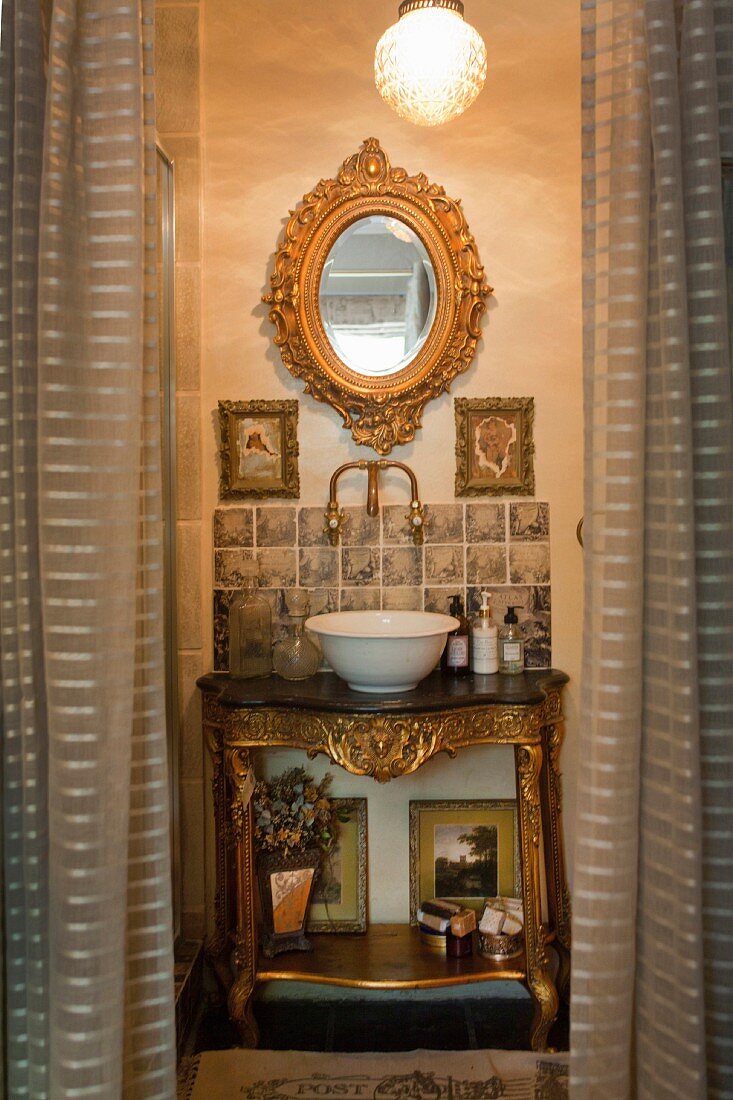 Baroque-style washstand with gilt details and white china basin below oval, gilt-framed mirror seen through open curtains in doorway