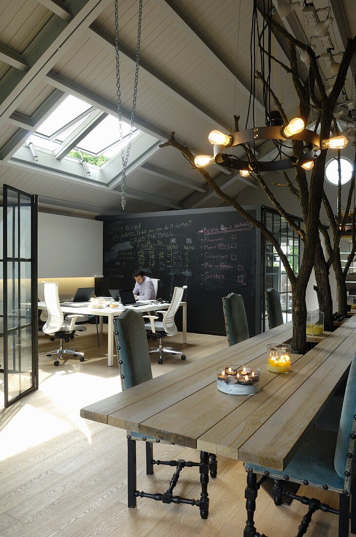 Blick über Esstisch mit integrierten Baumstämmen und Pendelleuchte aus schmiedeeisernem Ring auf Arbeitsplätze in loftarigem Raum