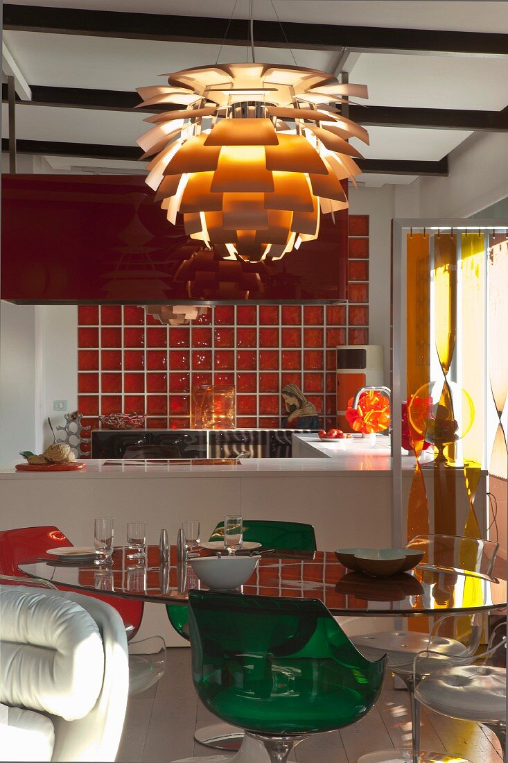 Dining table and coloured transparent plastic shell chairs below Artichoke pendant lamp; kitchen with orange glass tiles in background