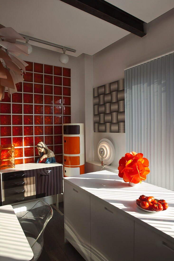 weiße Küchenzeile vor Fenster mit orangefarbener Tischleuchte und Retro-Sideboard mit Madonnenfigur vor orangeroter Glasfliesenwand