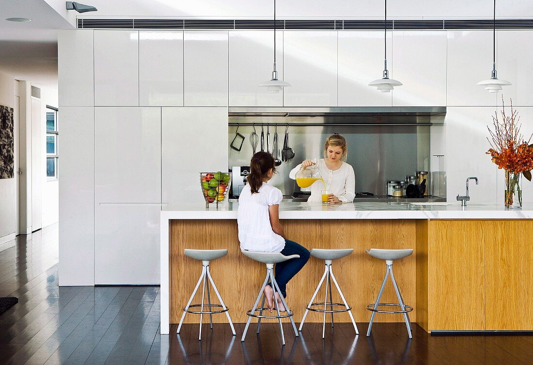 Two women at kitchen island with white worksurface and wooden front below classic pendant lamps