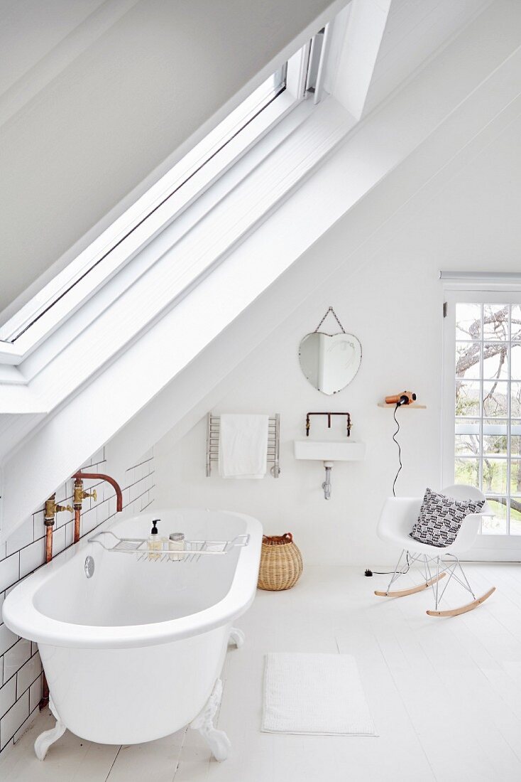 Free-standing vintage bathtub and classic rocking chair below sloping ceiling in white bathroom