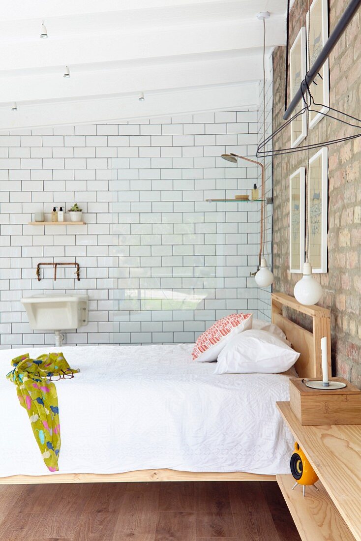 Doppelbett mit Holzgestell unter Pendelleuchten vor Ziegelwand, im Hintergrund Glasabtrennung mit Duschbereich und weißer Fliesenwand