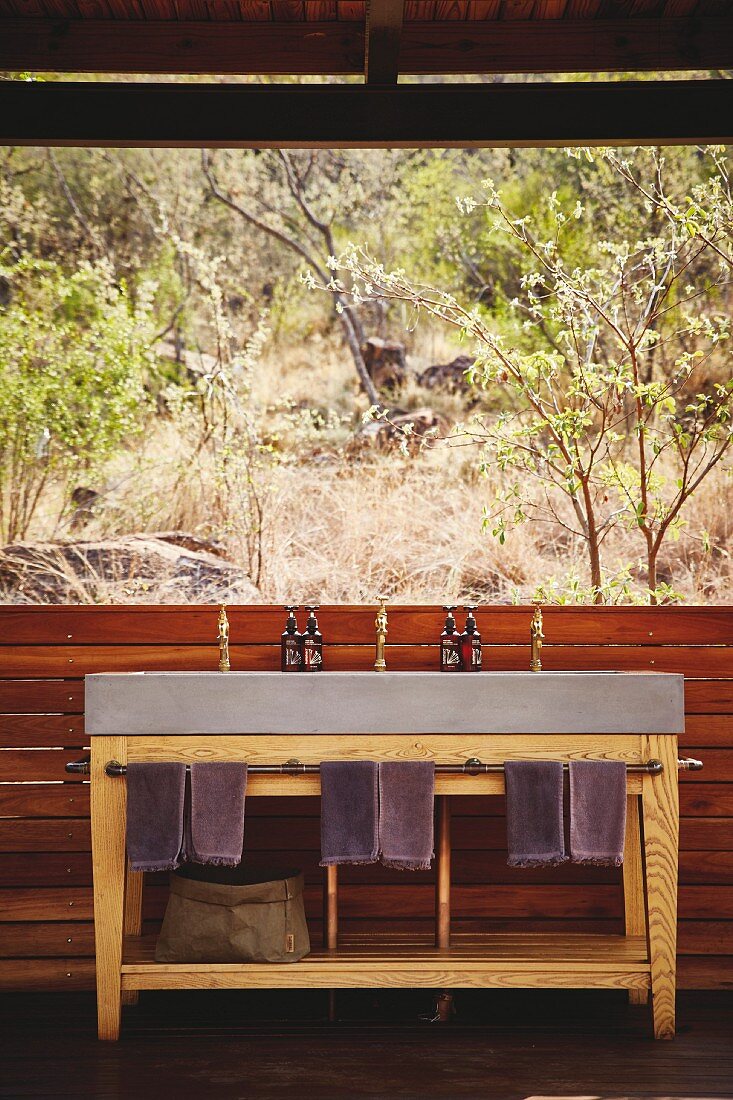 Beton-Waschtisch auf Holz-Untergestell mit aufgehängten Handtüchern vor Panoramafenster und Blick in Naturlandschaft