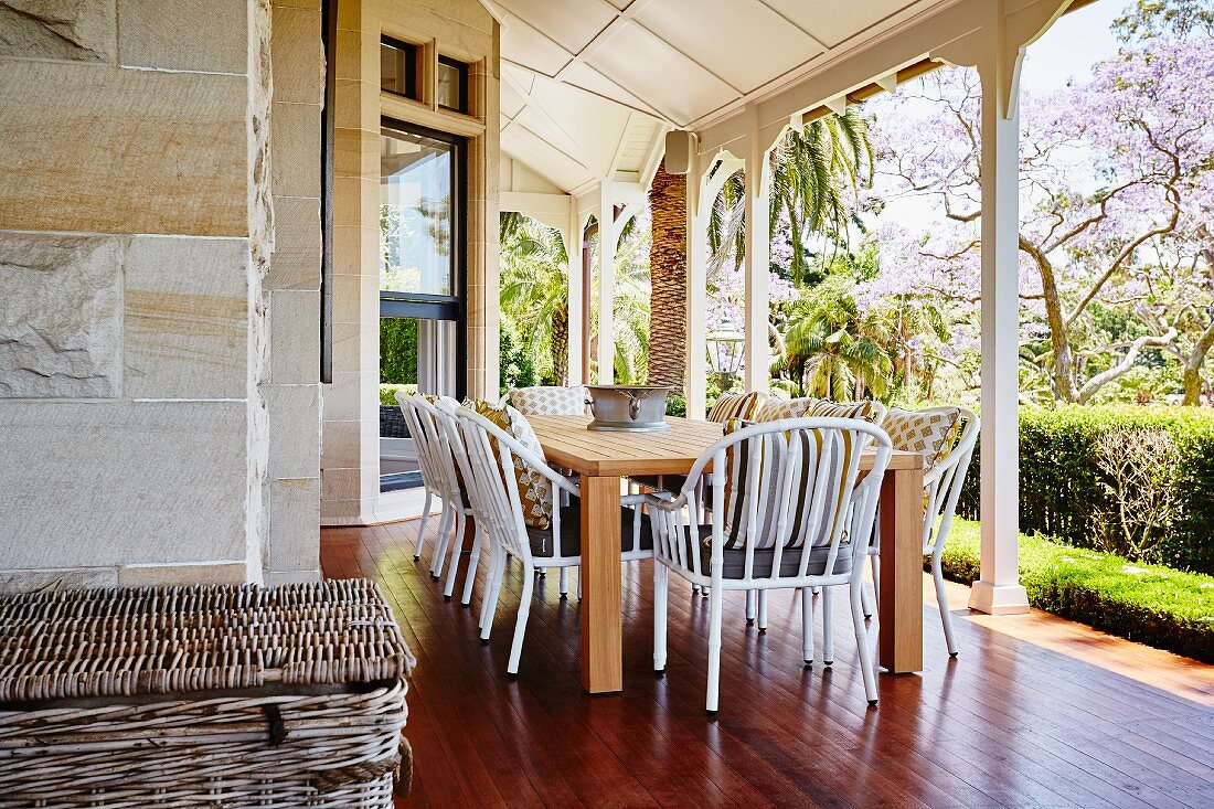 weiße Rattanstühle um Holztisch auf rötlichem Dielenboden einer Veranda mit weissen Holzstützen, vor Garten