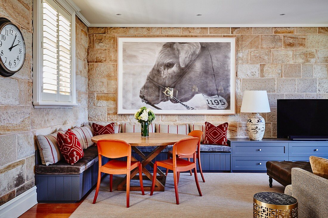 Gemütliche Wohnzimmerecke mit orangefarbenen Kunststoff Stühlen um Holztisch und blaue, eingebaute Eckbank an Natursteinwand