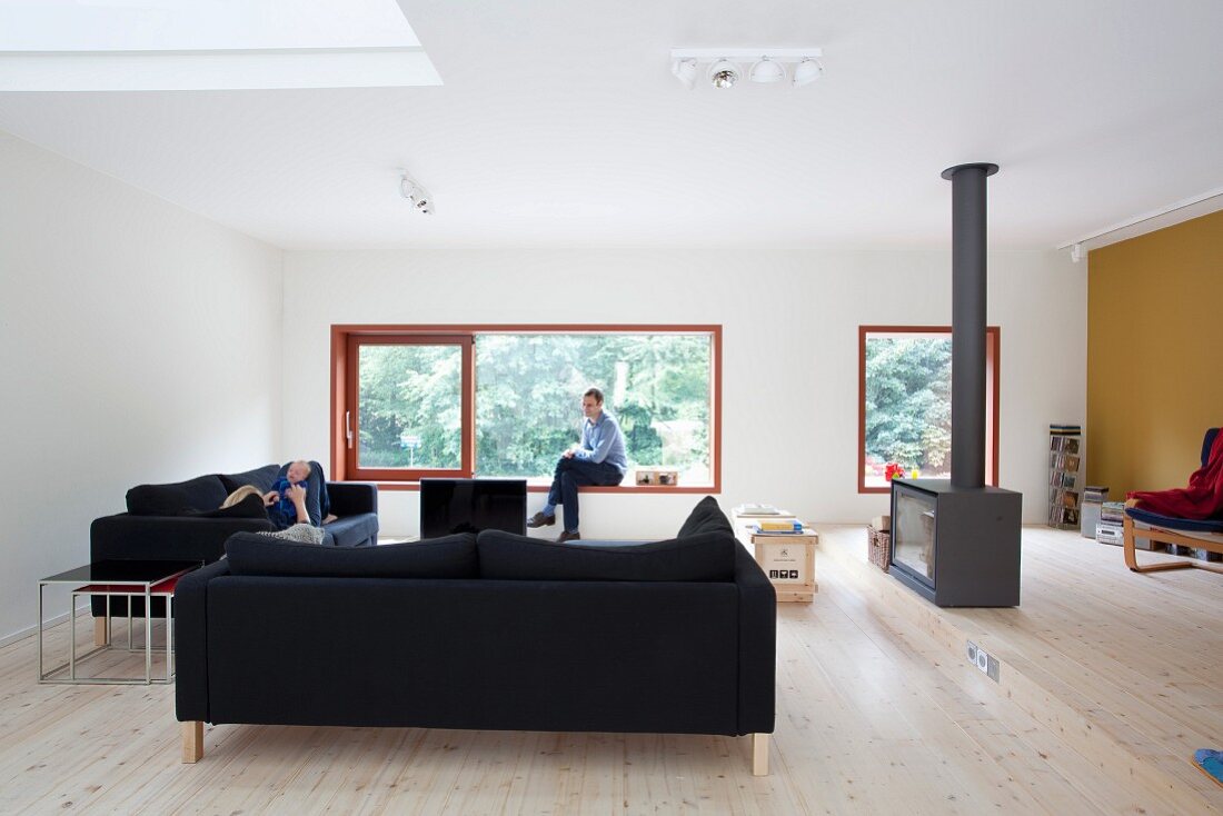 Loungeecke mit schwarzen Polstersofas und freistehender Kaminofen in minimalistischem Wohnraum, im Hintergrund Mann auf Fensterbank