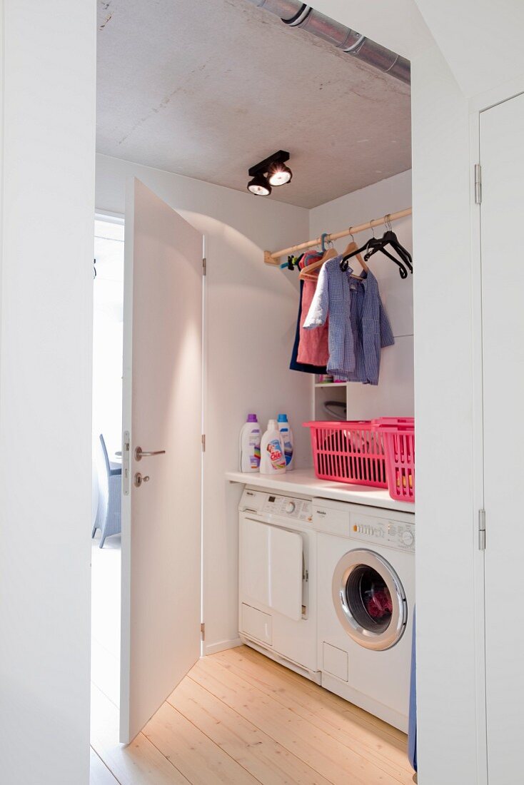 Blick in Waschraum auf Waschmaschine und Trockner unter Ablage, oberhalb Kleiderstange mit Kinderkleidung
