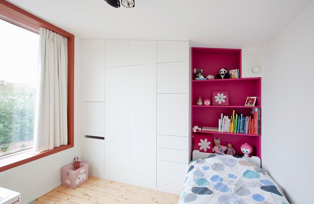 Kinderzimmer mit Bett vor pinkfarbenem Regal und weißem Einbauschrank