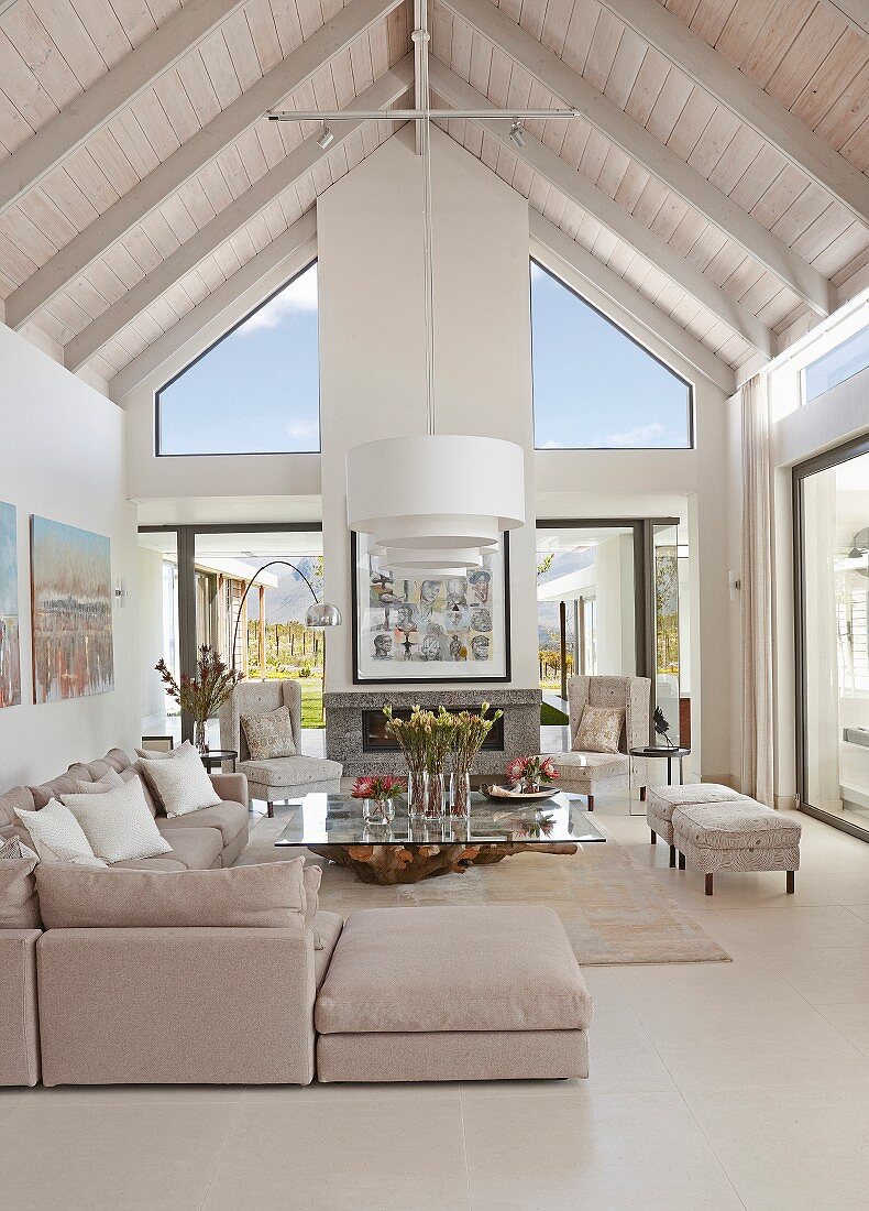 Offener, eleganter Wohnraum mit sichtbarem, weiß lasiertem Dachstuhl, hellgraues Übereck- Polstersofa und Glastisch