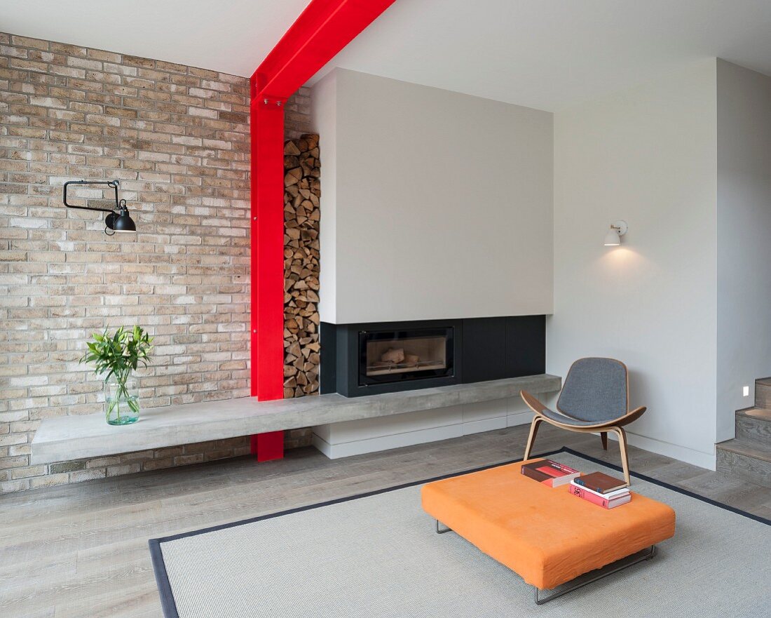Orangefarbener Bodentisch auf Teppich mit Designer-Sessel vor grauem Kamin, seitlich rote Stahlkonstruktion an Ziegelwand mit Brennholzlager