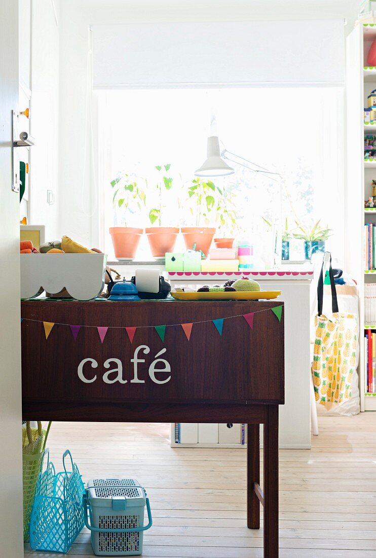 Sideboard decorated with bunting and lettering spelling 'café' in front of plants on windowsill in girl's bedroom