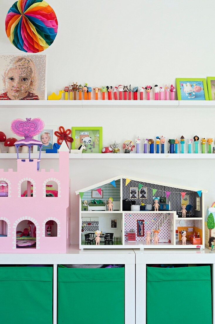 Puppenhaus und rosafarbenes Puppenschloss auf weißem Regalschrank mit grünen Aufbewahrungsboxen, darüber Spielzeugsammlung auf weißen Wandboards
