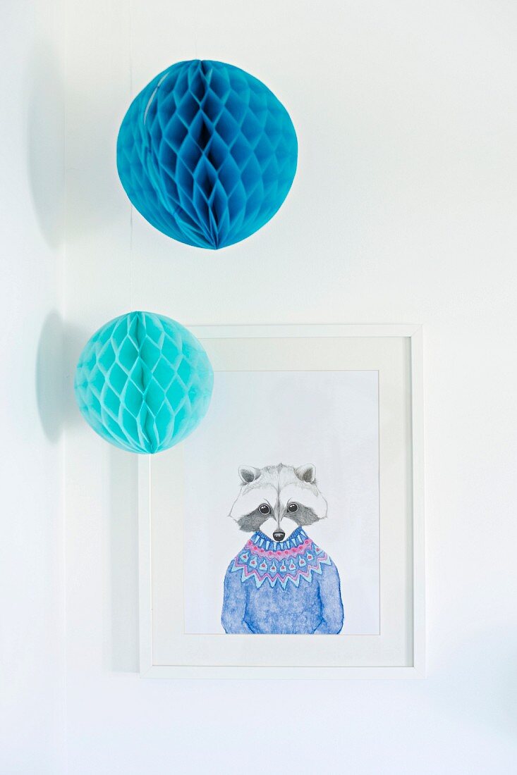 Two blue paper pompoms hung above white-framed drawing of racoon wearing jumper