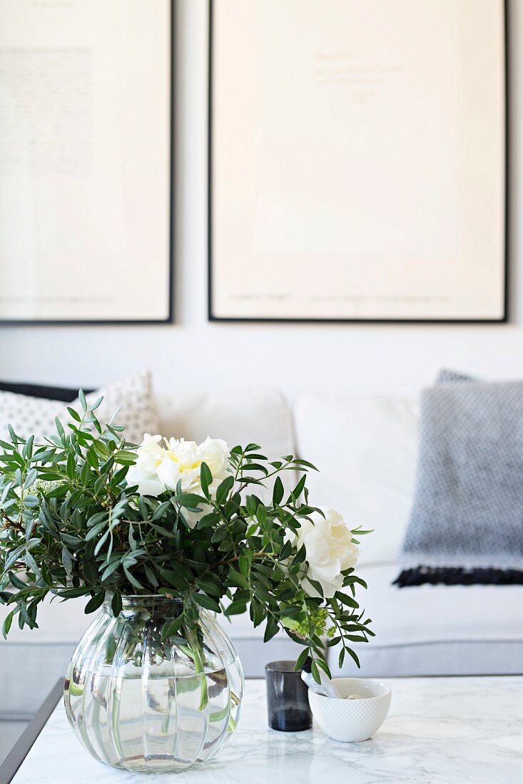 weiße Pfingstrosen mit Blätterzweigen in Glasvase auf Marmortisch vor Sofa