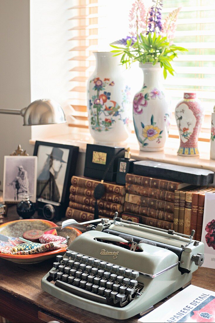 Vintage Schreibmaschine und Keramikschale vor antiquarischen Bücherstapeln auf Tisch am Fenster mit Vasen