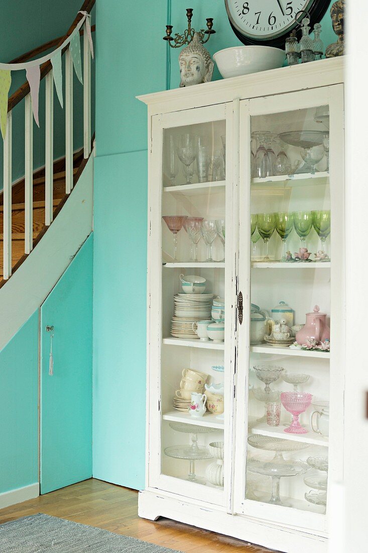 Crockery in vintage display case against turquoise wall next to wooden staircase with storage cupboards below