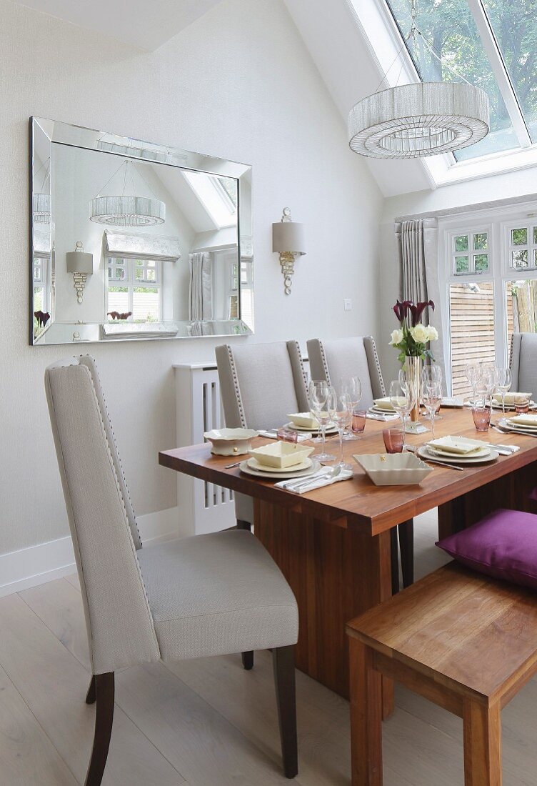 Elegant gedeckter Holztisch mit Sitzbank und hellgrauen Polsterstühlen, Wandspiegel und Oberlicht