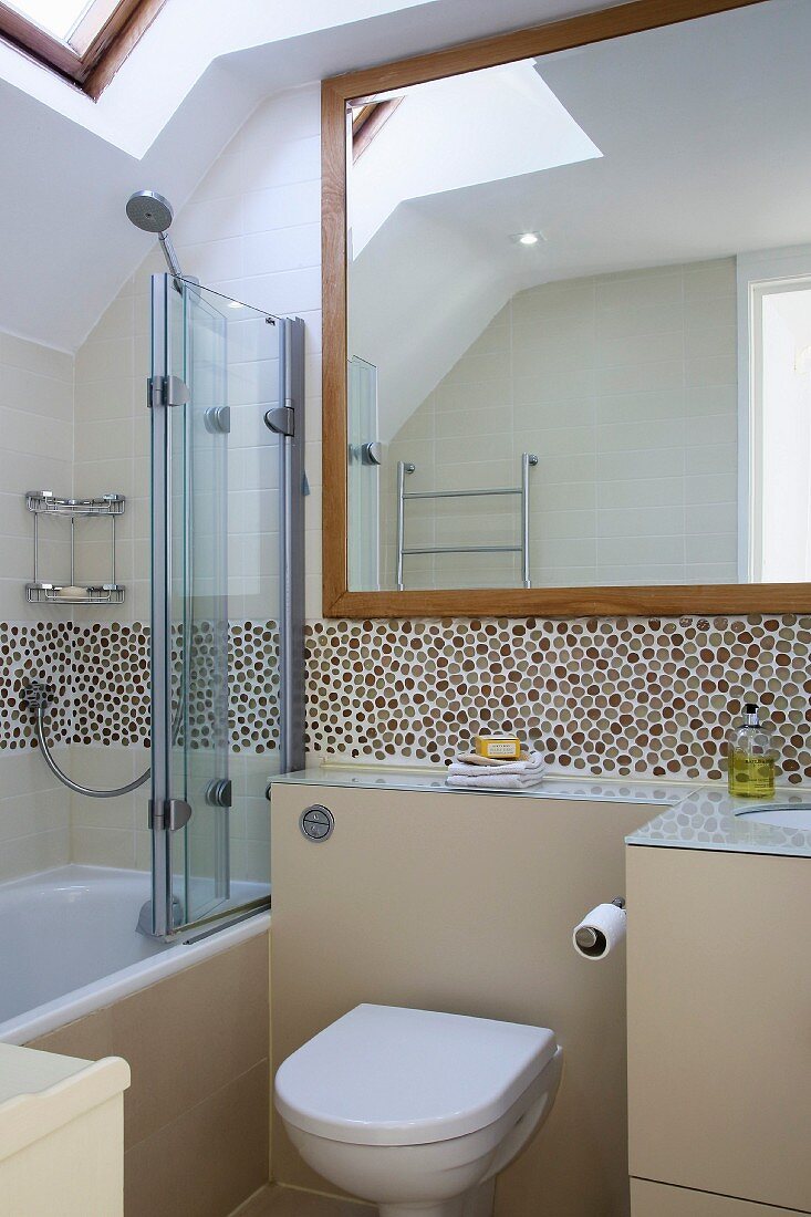 Badezimmer mit Toilette an hellbeiger Vormauerung, Mosaikfliesen an Wand, seitlich Badewanne mit Glasabtrennung