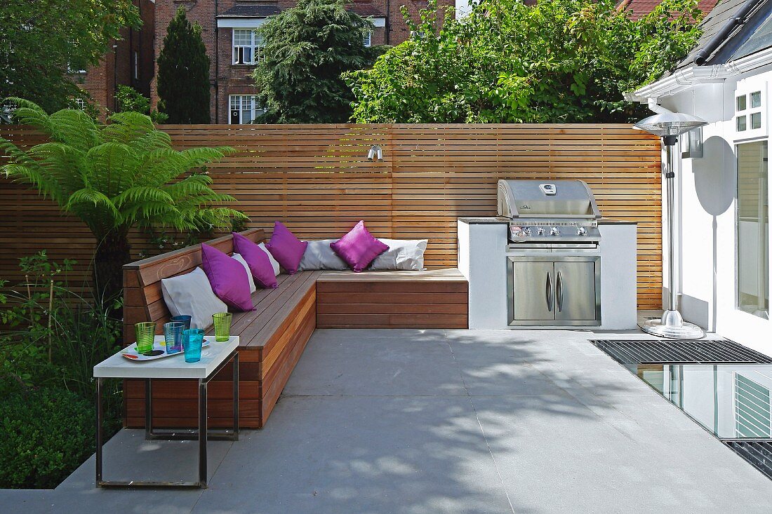 Eingebaute Holzbank mit violetten Kissen und Grill auf sommerlicher Terrasse, moderner Sichtschutzzaun