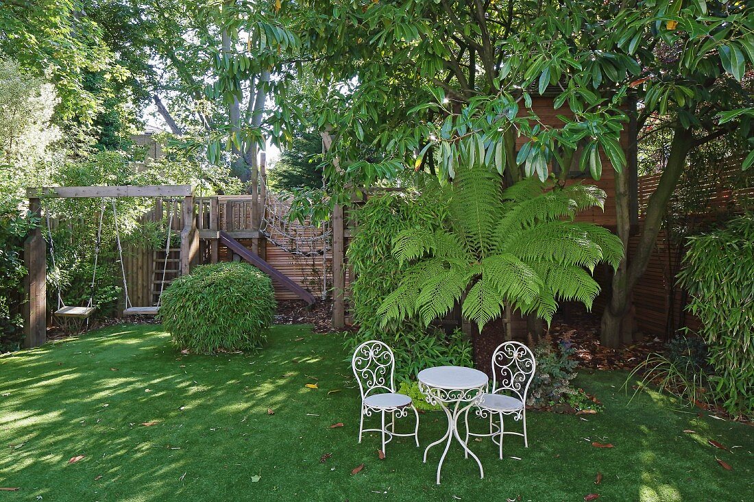 weiße Vintage Tisch mit Stühlen vor Palme und Bäumen im sommerlichen Garten, im Hintergrund Spielplatz mit Schaukelanlage