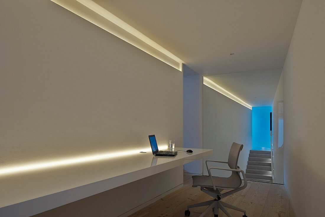 Minimalistisches Arbeitszimmer mit modernem Bürostuhl vor Theke mit indirekter Beleuchtung, im Hintergrund Treppenaufgang