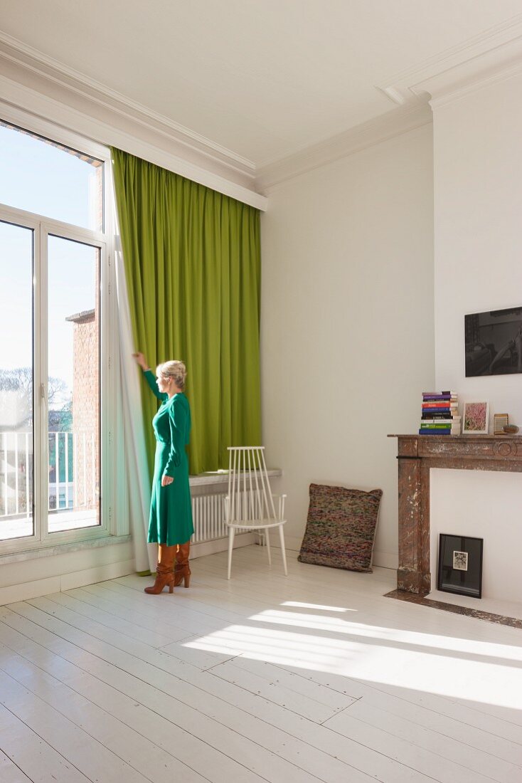 Woman standing by window with green curtain in period apartment with white wooden floor