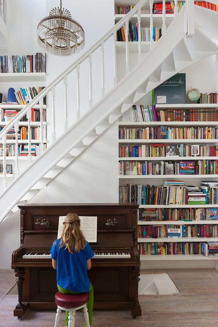 Girl playing piano at foot of staircase next to floor-to-ceiling bookcases