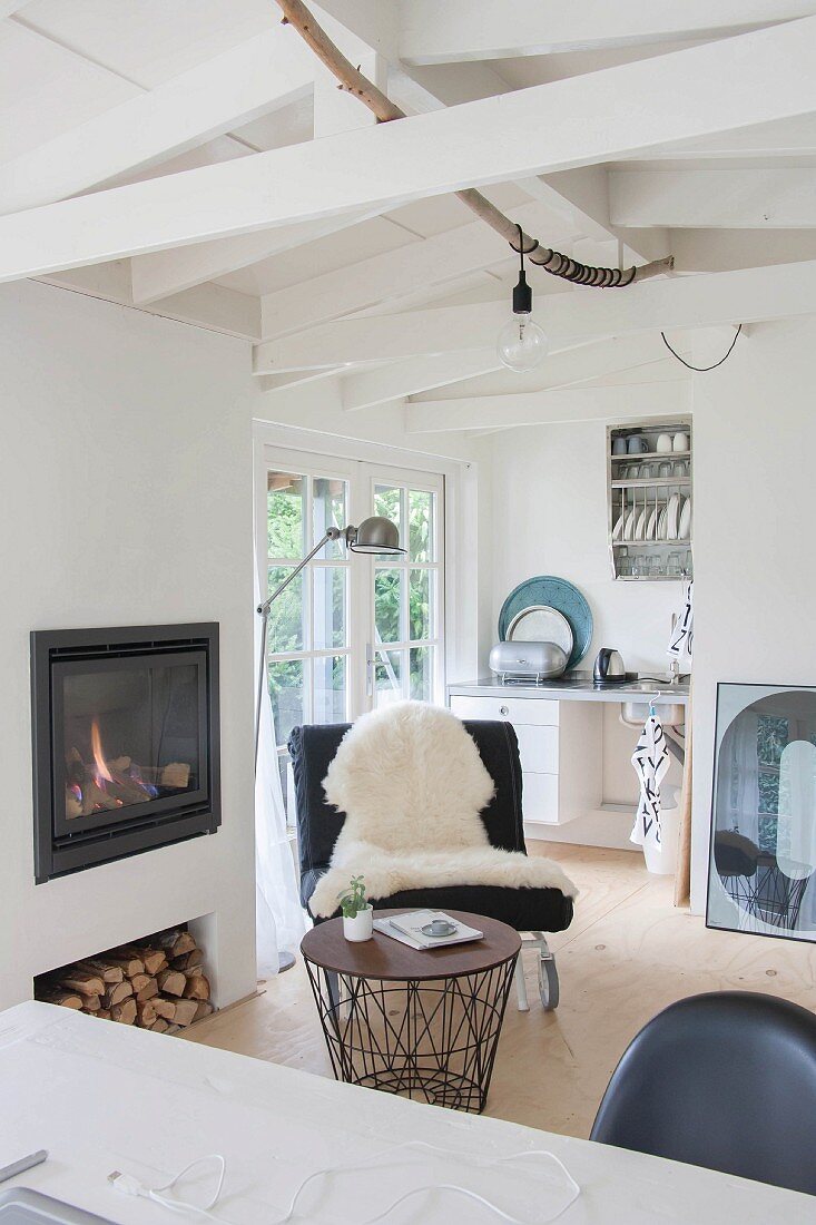 View across desk to black easy chair with white sheepskin and wire-framed side table in front of fireplace