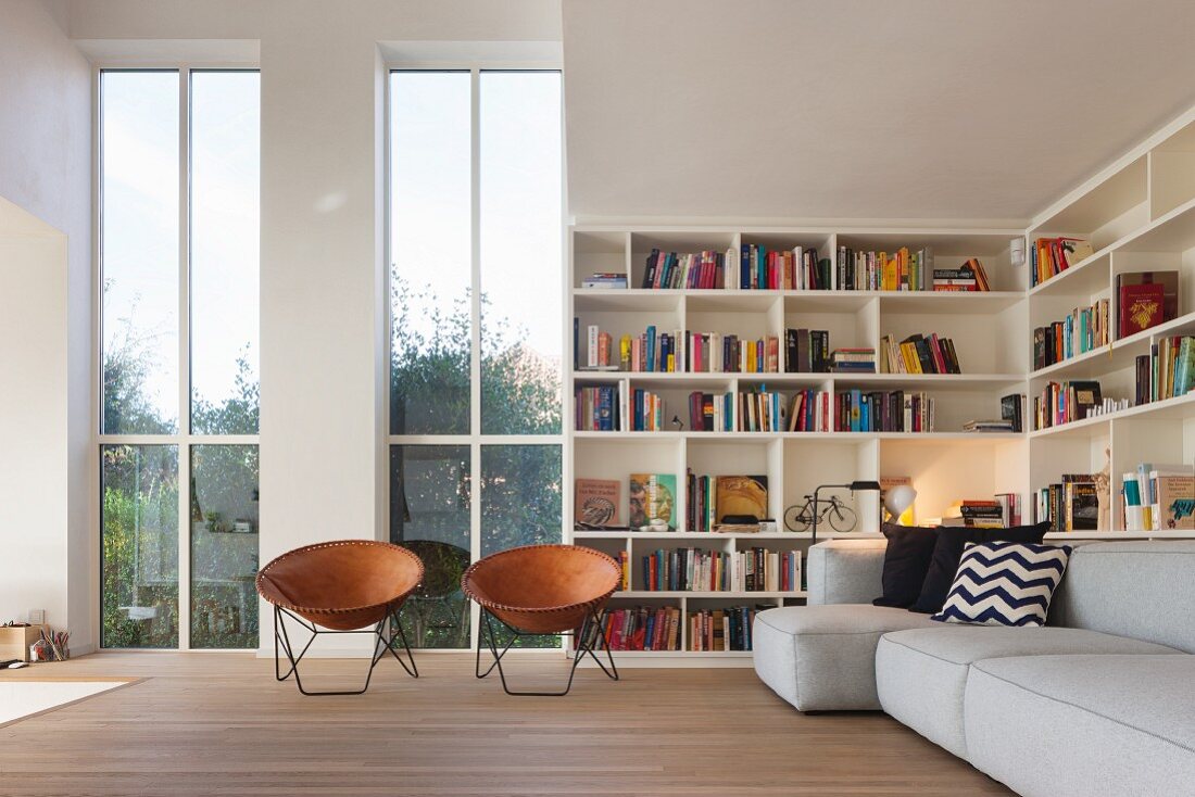 Retro-Ledersessel mit Schalensitz vor hohen Fenstern, seitlich Bücherregal und graues Polstersofa im Wohnzimmer