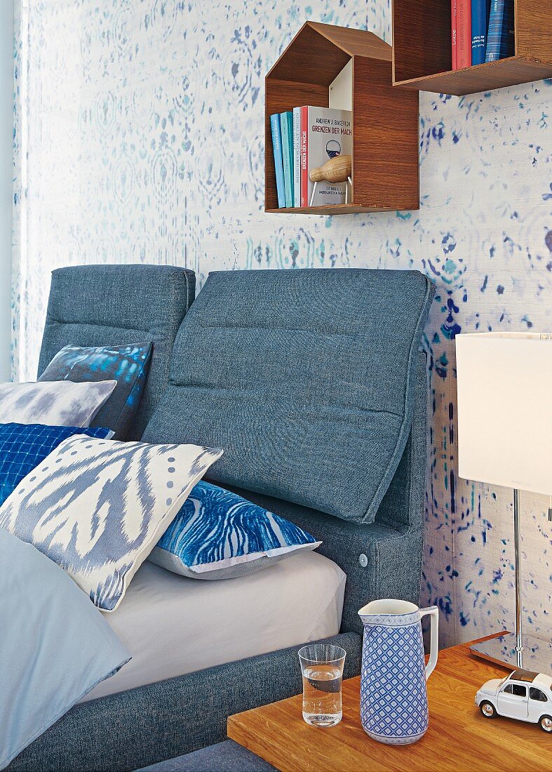 Double bed and headboard upholstered in denim below small, house-shaped shelving modules on wallpaper with blurry pattern